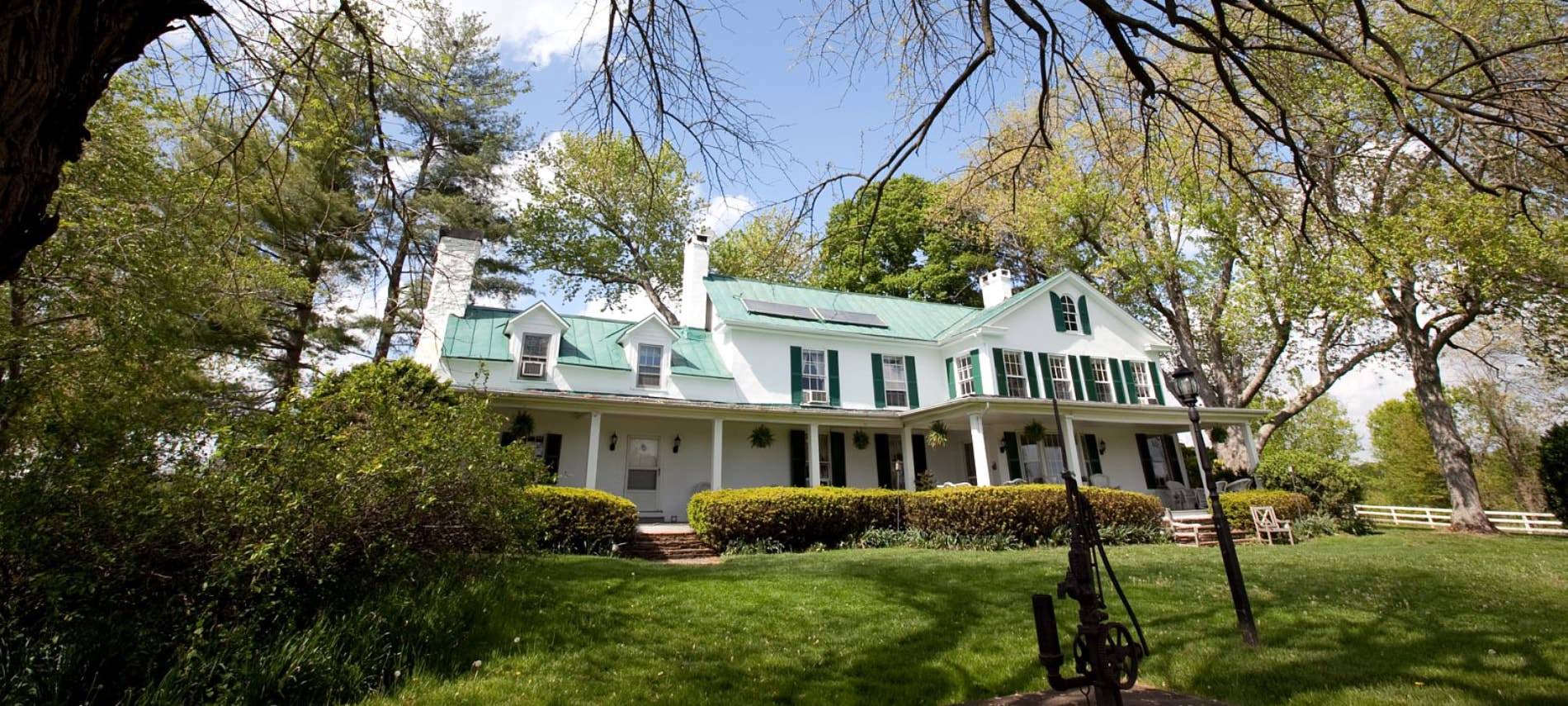 Exterior view of property painted white with dark shutters surrounded by a large green lawn, shrubs, and trees