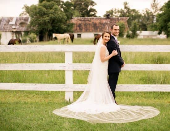Bride wearing white wedding dress and groom wearing black suite standing near white fence with old buildings and horses in the background