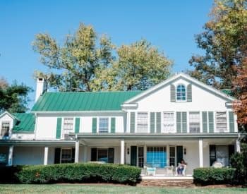 Exterior view of property painted white with green shutters and green roof surrounded by green shrubs and trees