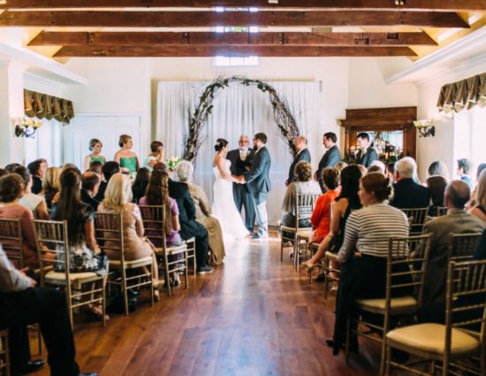 Large room with hardwood flooring and bride in white dress and groom in gray suit standing at the alter