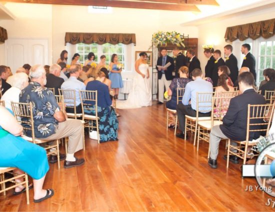 Large room with hardwood flooring and bride in white dress and groom in military uniform standing at the alter