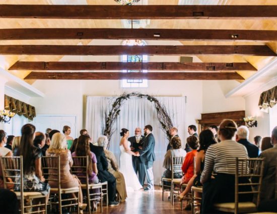 Large room with hardwood flooring and bride in white dress and groom in gray suit standing at the alter