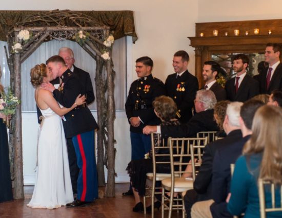 Bride in white dress and groom in military uniform kissing at the alter