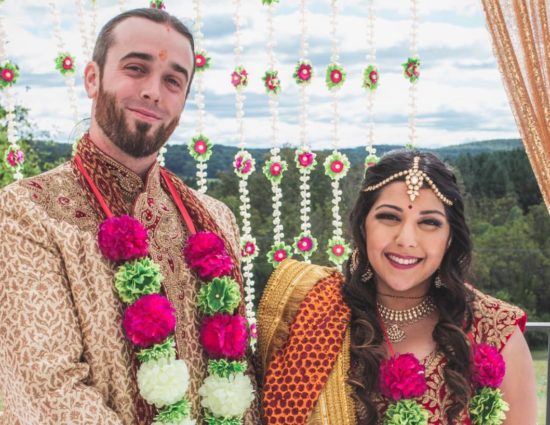 Bride and groom dressed in traditional Indian wedding attire with white, green, and pink flower leis