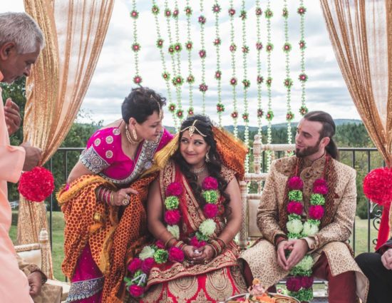 Bride and groom dressed in traditional Indian wedding attire with white, green, and pink flower leis with family nearby