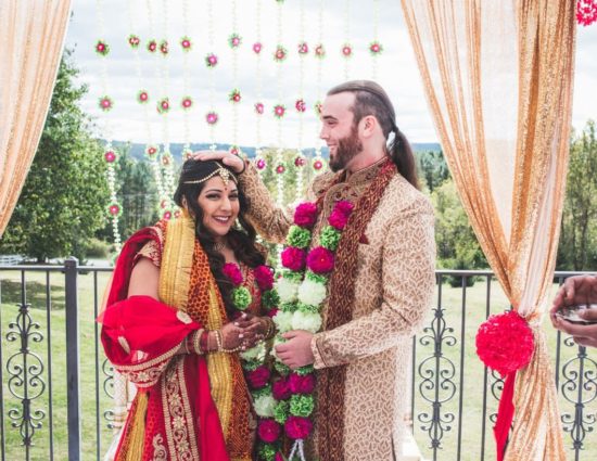 Bride and groom dressed in traditional Indian wedding attire with white, green, and pink flower leis