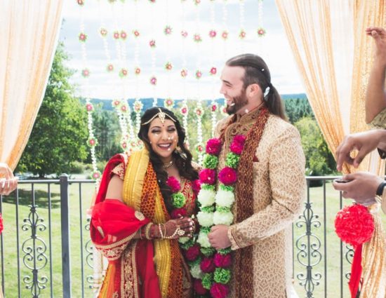 Bride and groom dressed in traditional Indian wedding attire with white, green, and pink flower leis