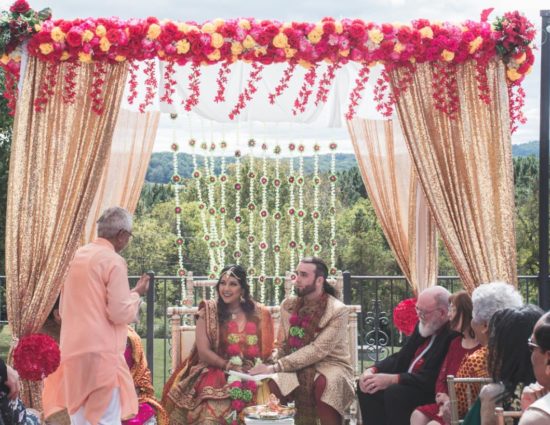Bride and groom dressed in traditional Indian wedding attire with white, green, and pink flower leis with family nearby