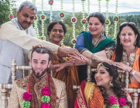 Bride and groom dressed in traditional Indian wedding attire with white, green, and pink flower leis and family members standing behind with hands on their heads