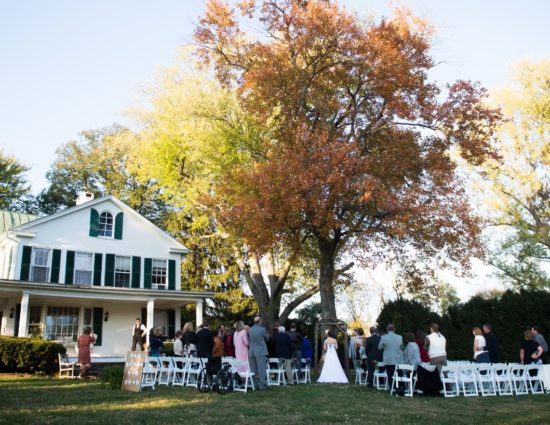 Wedding ceremony outside front of white house near large tree with bride in white dress and groom in gray suit