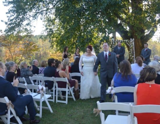 Wedding ceremony outside near large tree with bride in white dress and groom in gray suit walking down the aisle