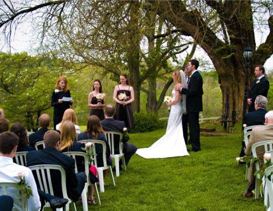 Wedding ceremony outside near large tree with bride in white dress and groom in black suit