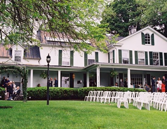 Exterior view of main house painted white with green shutters with lawn set up for wedding ceremony with white chairs