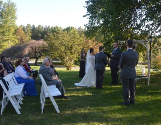 Wedding ceremony outside near large tree with bride in white dress and groom in gray suit