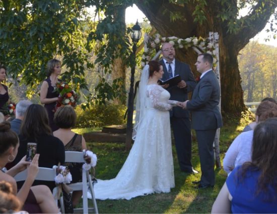 Wedding ceremony outside near large tree with bride in white dress and groom in gray suit