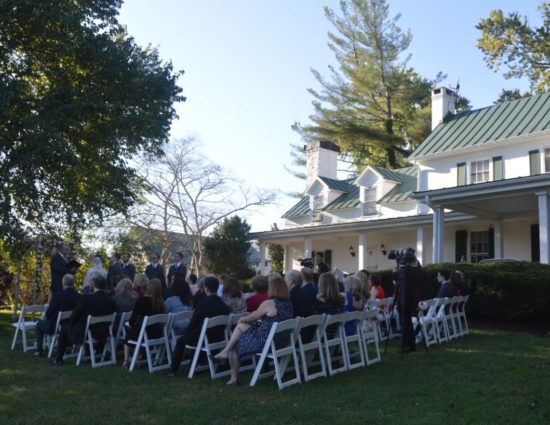 Wedding ceremony outside near large tree and front of house with bride in white dress and groom in gray suit