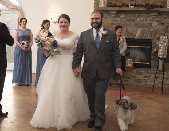 Large room with concrete flooring, bride in white dress, groom in gray suit, and dog walking down the aisle