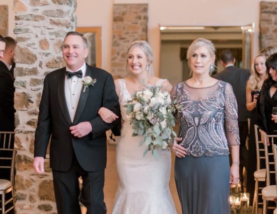 Bride in white dress with father in black suit and mother in gray dress walking bride down the aisle