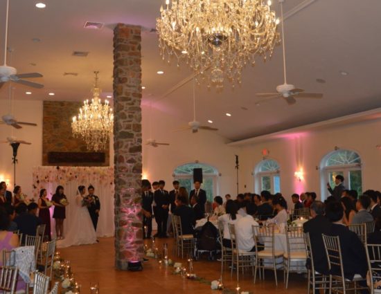 Large room with concrete flooring, bride in white dress and groom in black suit standing at the alter, and people sitting at tables watching the ceremony