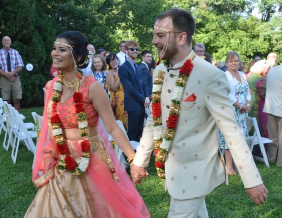 Bride and groom in traditional Indian wedding attire walking down aisle together holding hands