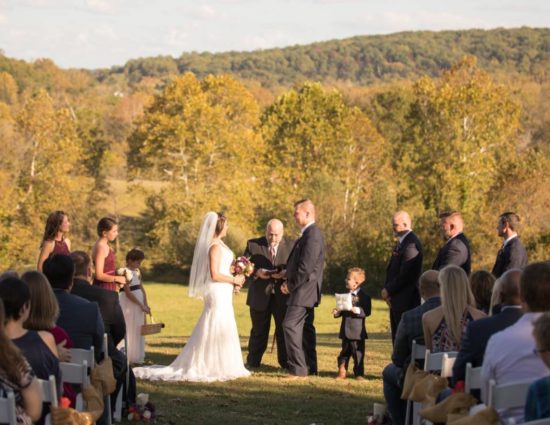 Wedding ceremony in front of rolling hills with green trees with bride in white dress and groom in black suit