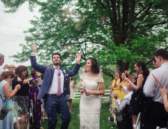 Wedding ceremony in front of large tree with green leaves with bride in white dress and groom in dark purple suit walking down the aisle