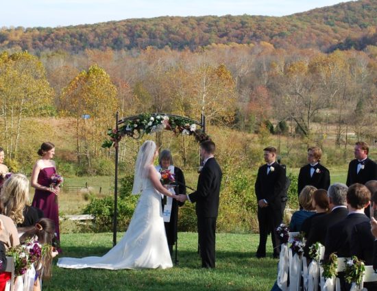 Wedding ceremony with rolling hills of trees with fall color in the background with bride in white dress and groom in black suit