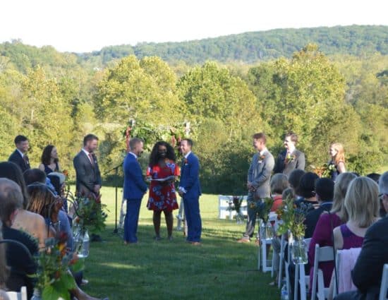 Wedding ceremony in front of rolling hills with green trees with two grooms in blue suits standing at alter