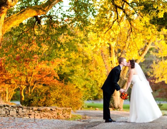 Bride with white dress and groom with black suit standing on gravel road surrounded by trees with fall color