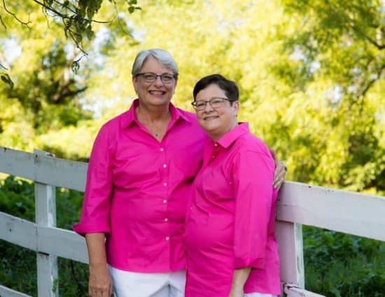 Two brides with pink shirts and white pants standing by white fence