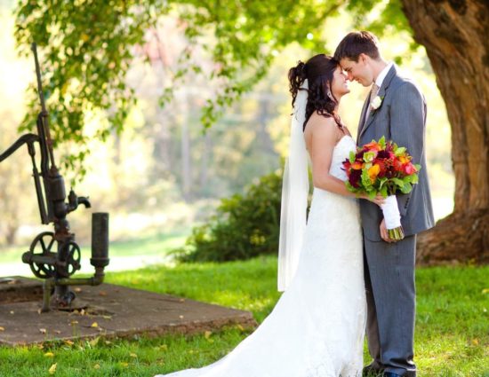 Bride with white dress and groom with gray suit standing by large tree and old water pump