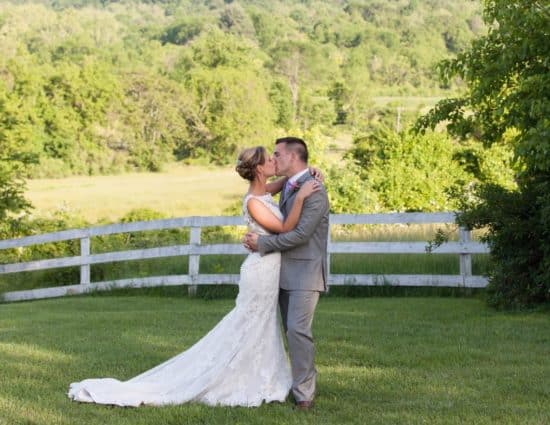 Bride with white dress and groom with gray suit standing by white fence with trees in the background