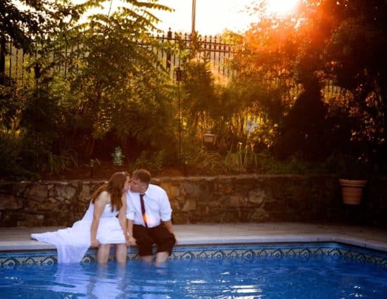 Bride with white dress and groom with white shirt and black pants sitting by pool with legs in the water