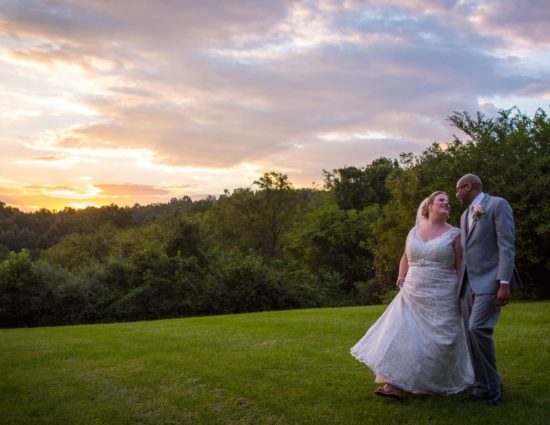 Bride with white dress and groom with gray suit standing on green grass with the sun setting behind them