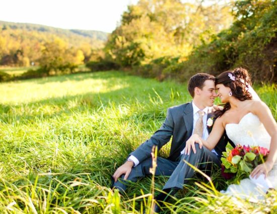 Bride with white dress and groom with gray suit sitting in field of green grass