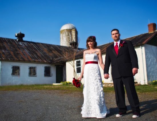 Bride with white dress and groom with black suit standing in front of old barn and silo