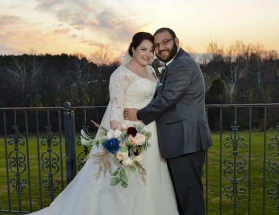 Bride with white dress and groom with gray suit standing by rod iron railing as sun sets