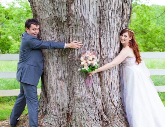 Bride with white dress and groom with gray suit hugging large tree