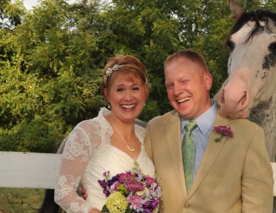 Bride with white dress and groom with tan suit standing by white and black horse with head on groom's shoulder