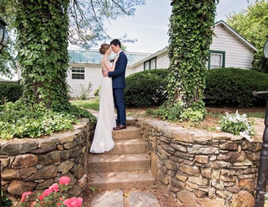 Bride with white dress and groom with navy suit standing on steps in front of white cottage