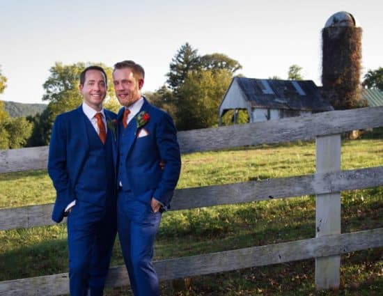 Two grooms in blue suits standing by old fence with old barn and silo in background