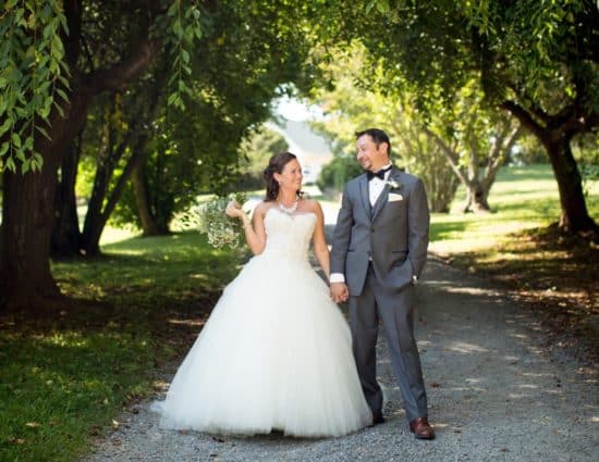 Bride with white dress and groom with gray suit standing on gravel road surrounded by large trees with green leaves