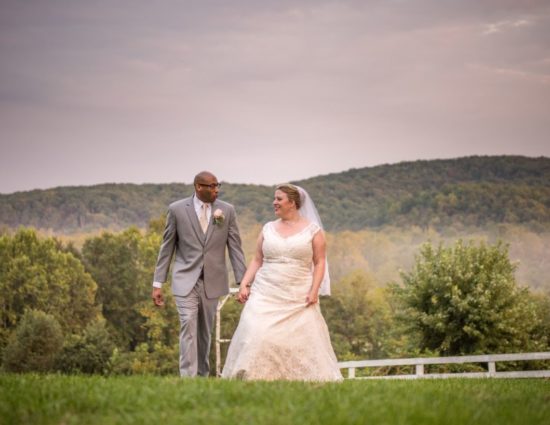 Bride with white dress and groom with gray suit standing near white fence with rolling hills in the background