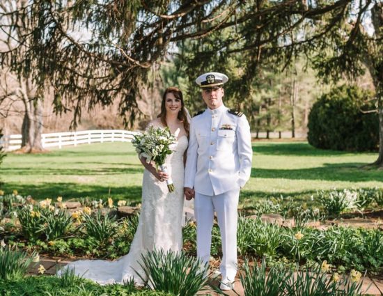 Bride with white dress and groom with white naval uniform standing on stone path surrounded by white and yellow flowers