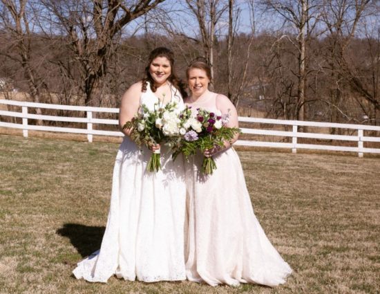 Two brides with white dresses standing in grass with white fence behind them