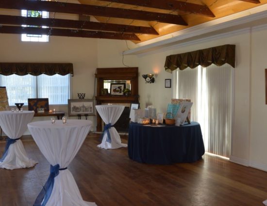 Large room with hardwood flooring set up for a cocktail reception with tables and white tablecloths and one table with blue tablecloth