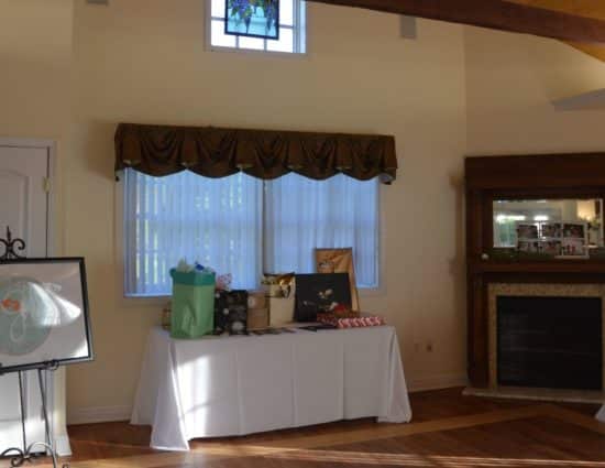 Large room with hardwood flooring, fireplace in the corner, long table with white tablecloth and wedding gifts, and cocktail table with white tablecloth