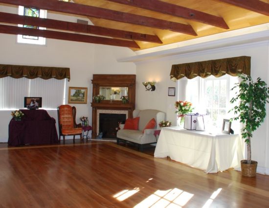 Large room with hardwood flooring, long table with white tablecloth, an upholstered chair, an upholstered love seat, and fireplace in the corner