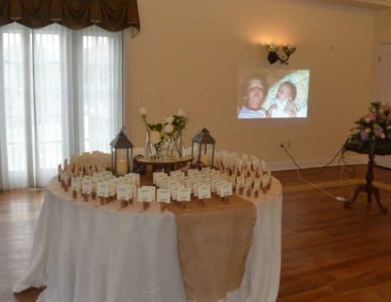 Large room with hardwood flooring, large round table with white tablecloth and wedding party favors, and slideshow in the background