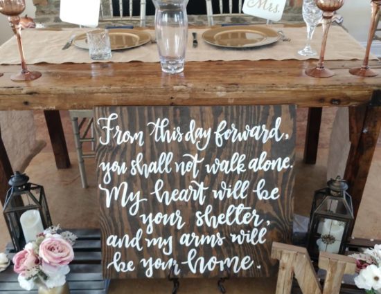 Old wooden table set up for bride and groom, a wooden panel with a saying in white lettering, and white and pink flowers in vases